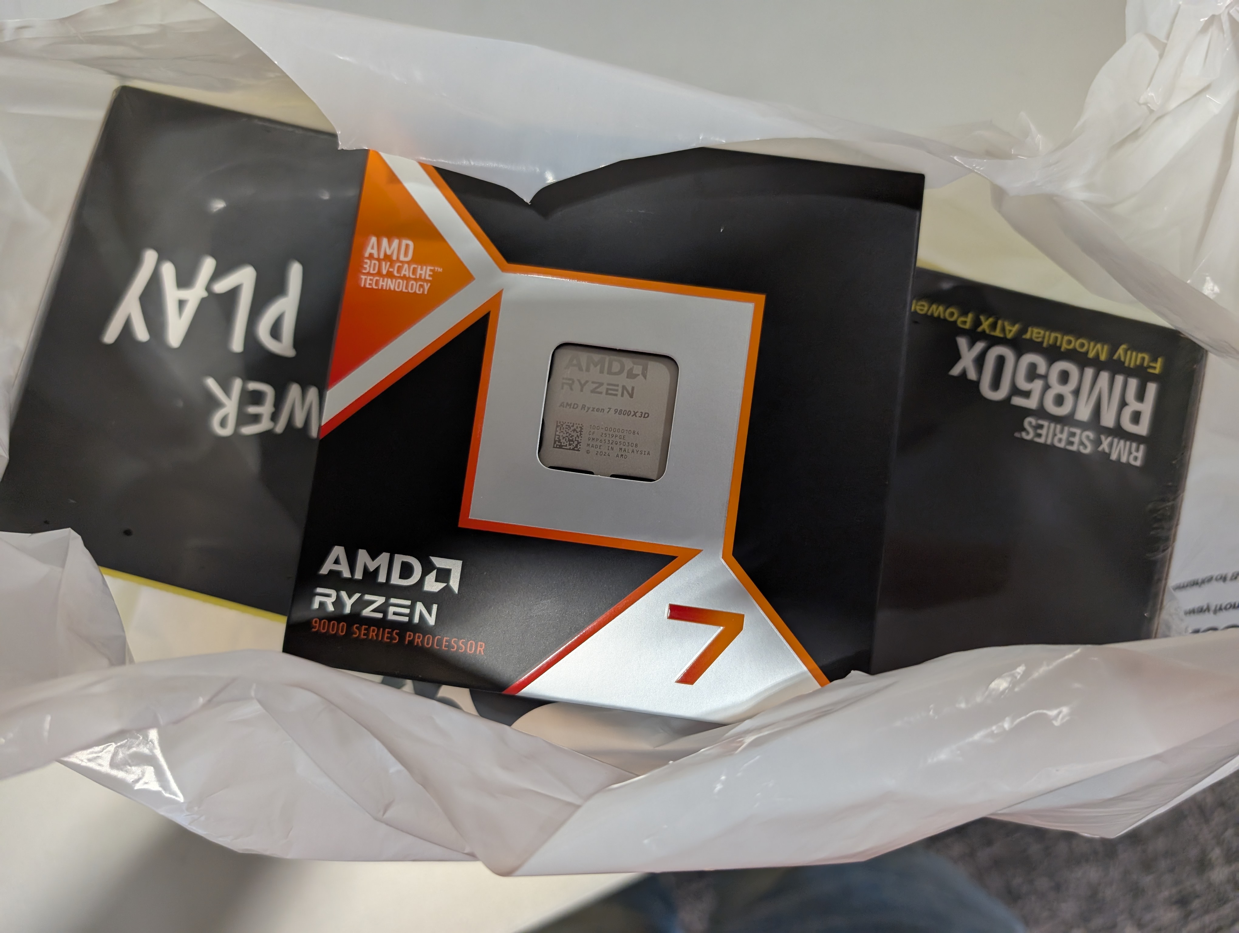 The picture shows an image of the Ryzen 7 9800X3D CPU on top of a Corsair RM850X power supply inside a BestBuy bag.